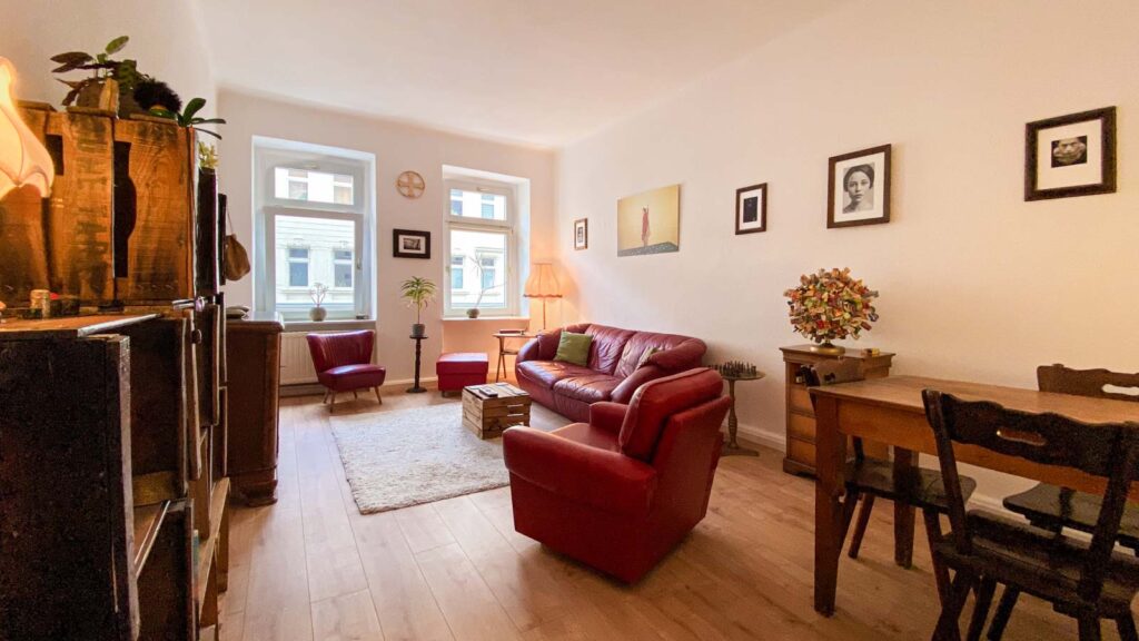 Helles Zimmer mit Holzboden, roten Ledermöbeln, einem Teppich, verschiedenen Pflanzen, einer Stehlampe, mehreren gerahmten Bildern an der Wand und einem Holztisch mit Stühlen.