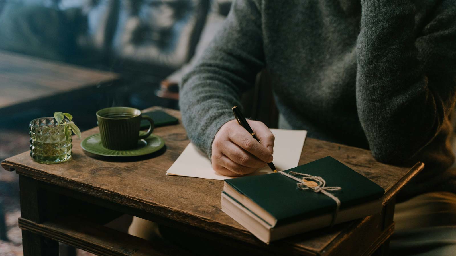 Person in grauem Pullover schreibt mit einem Stift auf ein Blatt Papier an einem rustikalen Holztisch, neben einer gebundenen grünen Mappe, einer grünen Tasse mit Kaffee und einem Glastopf mit einer kleinen Pflanze.