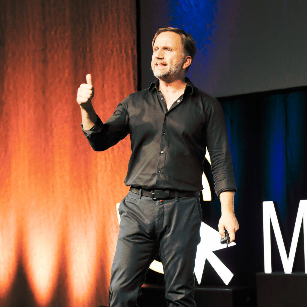 Ein Mann in schwarzem Hemd und dunkler Hose steht auf einer Bühne vor einem orangen und schwarzen Hintergrund und gibt mit seiner linken Hand einen Daumen nach oben, während er einen Präsentationsklicker in der rechten Hand hält.