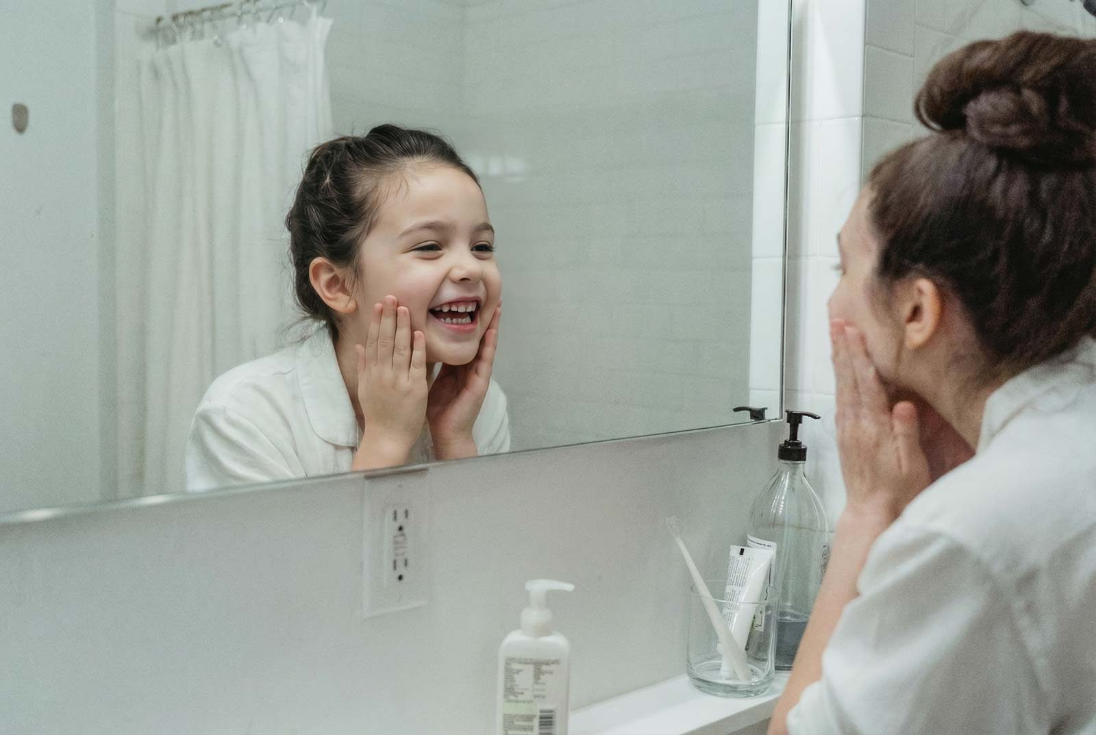 Ein Frau betrachtet sich im Badezimmerspiegel und berührt mit beiden Händen ihre Wangen. Im Spiegelbild zeigt sich ihr fünfjähriges Ich.